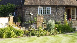 A stone house with a sloped roof, a garden with various plants and flowers, and a lawn in front. - Magpies (Horsham)
