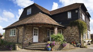 A two-story stone house with a steeply pitched roof, a front porch with steps, and a small garden with potted plants. - Magpies (Horsham)