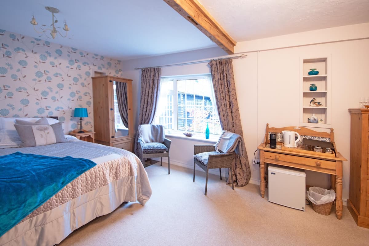 A bedroom with a bed, a desk, a chair, a small fridge, and a window with curtains.