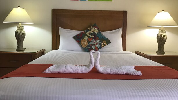 A neatly made bed with a red bedspread, two matching lamps on either side, and a colorful pillow on the headboard. - Alicia's Inn (Philipsburg)