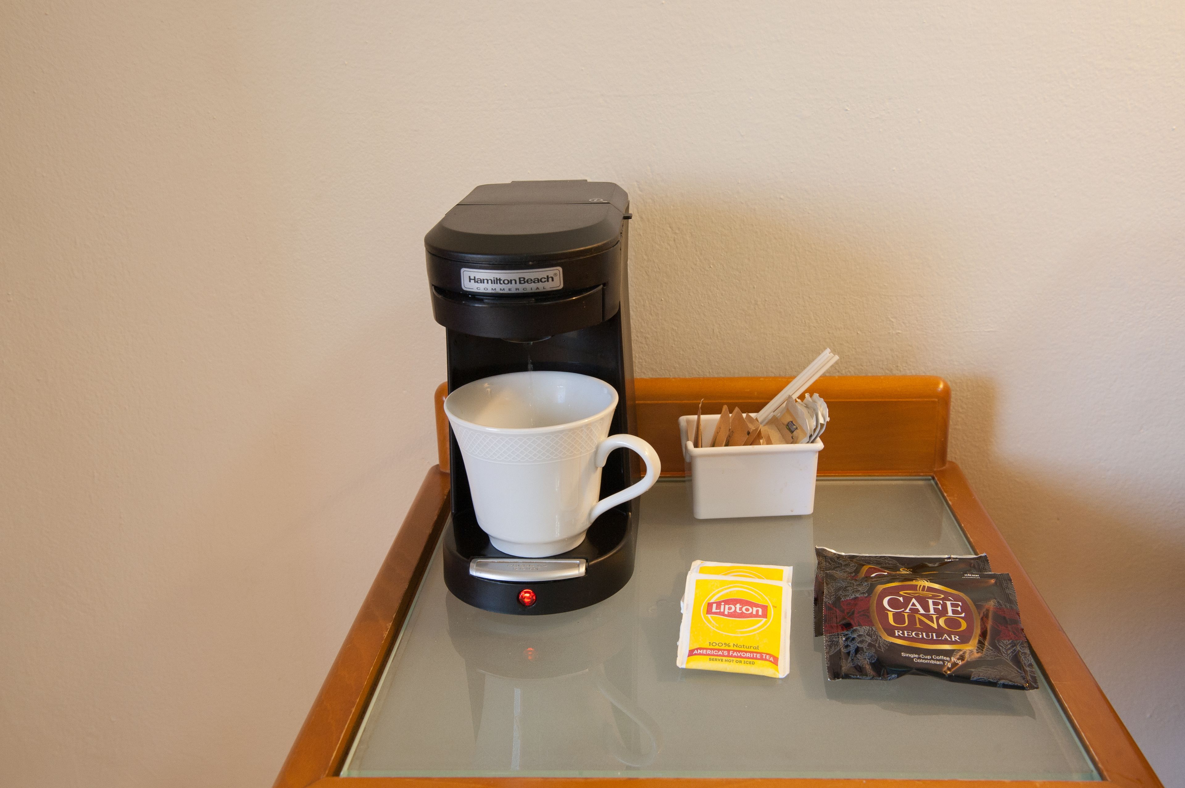 Une machine à café, une tasse, un récipient contenant des ustensiles et une boîte de thé Lipton sur une table en verre.