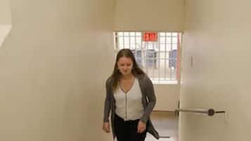 Una persona bajando una escalera con barandas rojas en un edificio de paredes lisas y una ventana con un letrero de salida.
