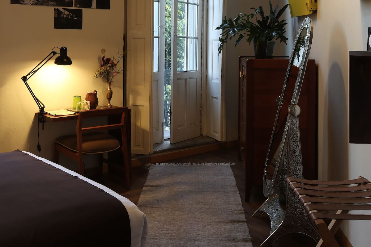 A bedroom with a bed, desk, lamp, chair, and a large window.