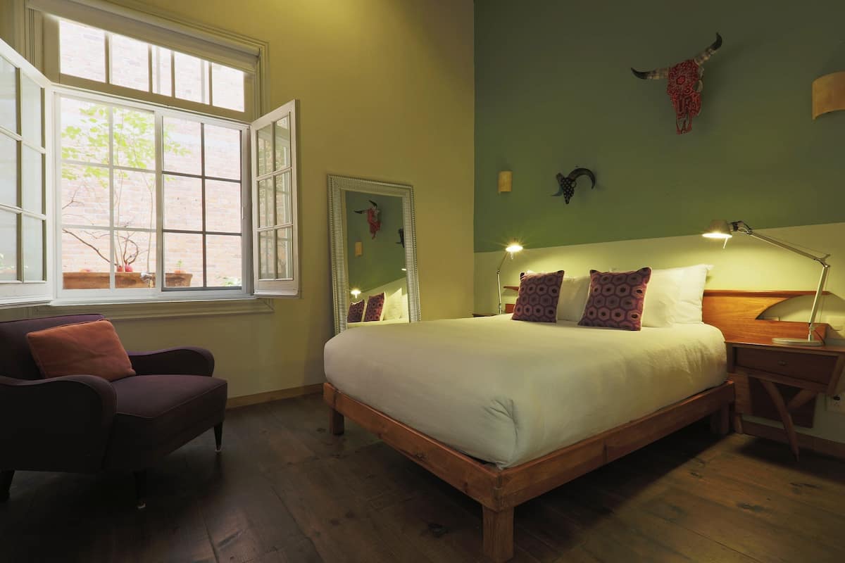 A bedroom with a large bed, a chair, a nightstand, a lamp, and a window with a view of a tree.