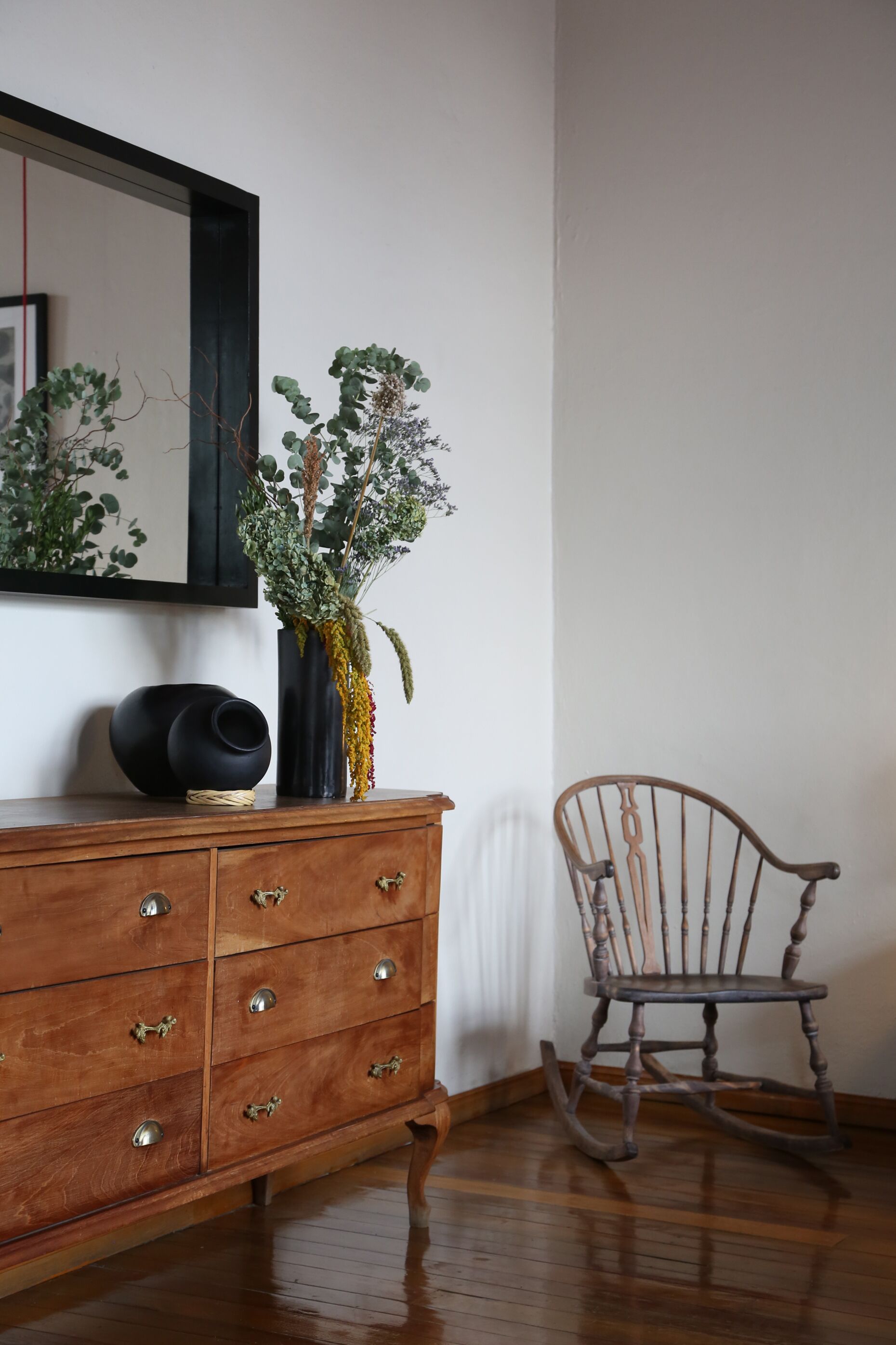 Un aparador de madera con cajones, un florero negro con plantas, una mecedora y un espejo en la pared.