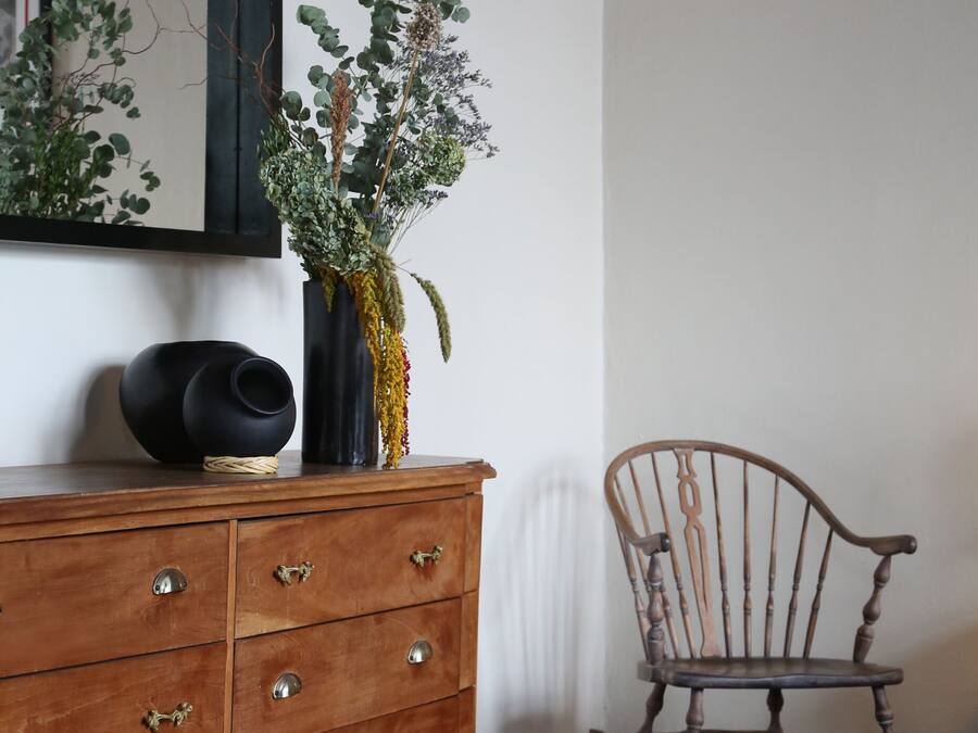 Un aparador de madera con cajones, un florero negro con plantas, una mecedora y un espejo en la pared.