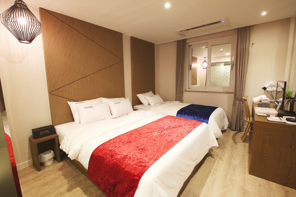 A hotel room with two beds, a desk, a chair, a small table, a mirror, and a window with curtains.