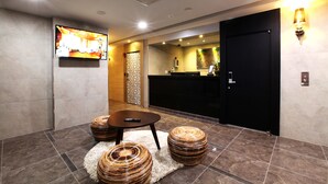 A modern hotel lobby with a reception desk, a television mounted on the wall, and a seating area with a round table and ottomans. - Jeju Lux Hotel (Seogwipo)