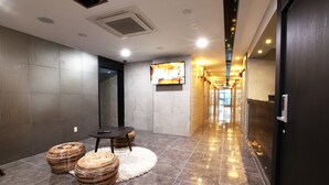 A modern hotel lobby with a tiled floor, a round table with stools, and a flat-screen TV mounted on the wall. - Jeju Lux Hotel (Seogwipo)