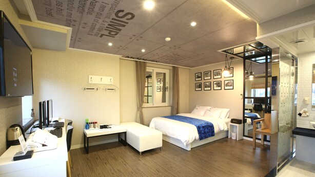 A modern hotel room with a bed, bedside table, desk, and a bathroom with a mirror and sink. - Jeju Lux Hotel (Seogwipo)