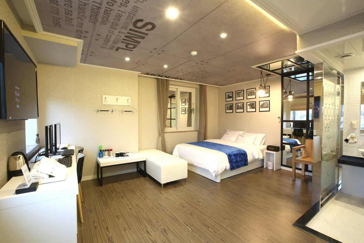 A modern hotel room with a bed, bedside table, desk, and a bathroom with a mirror and sink.