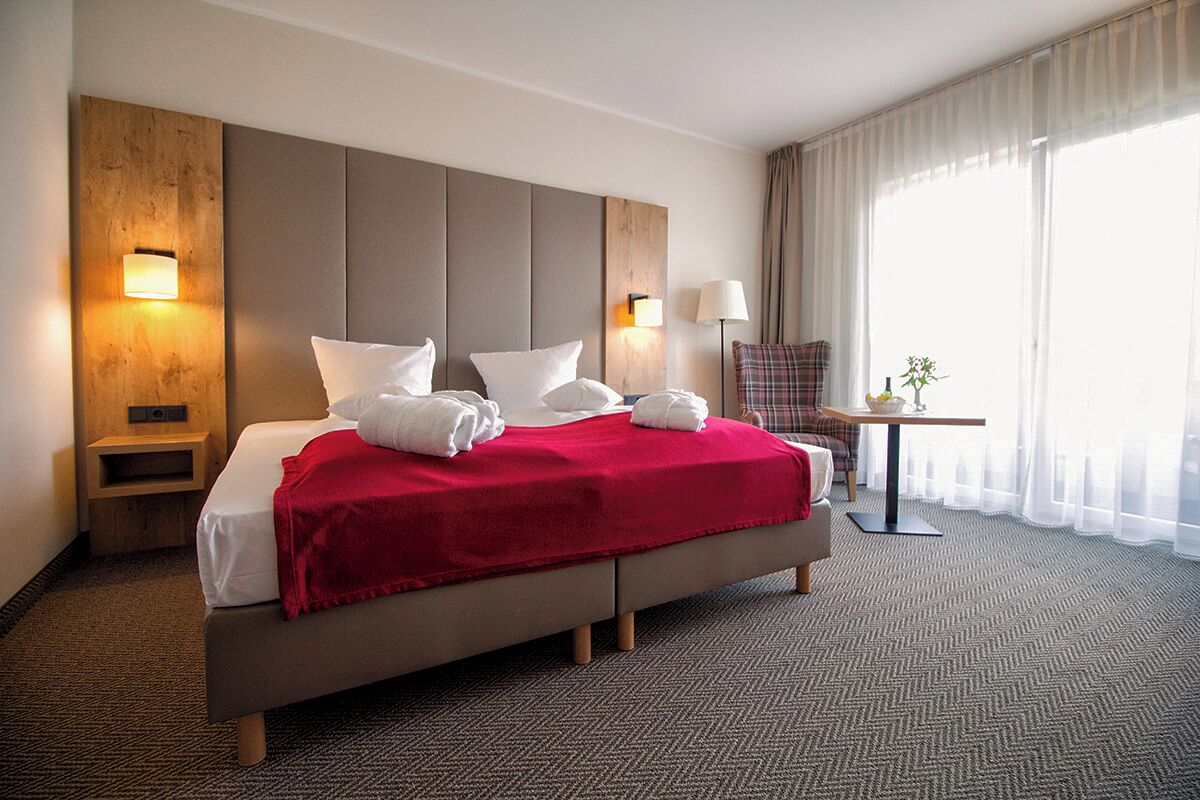 A hotel room with a large bed, two bedside tables, a small table with a lamp, and a view of the outdoors through sheer curtains.