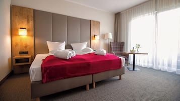 A hotel room with a large bed, two bedside tables, a small table with a lamp, and a view of the outdoors through sheer curtains.