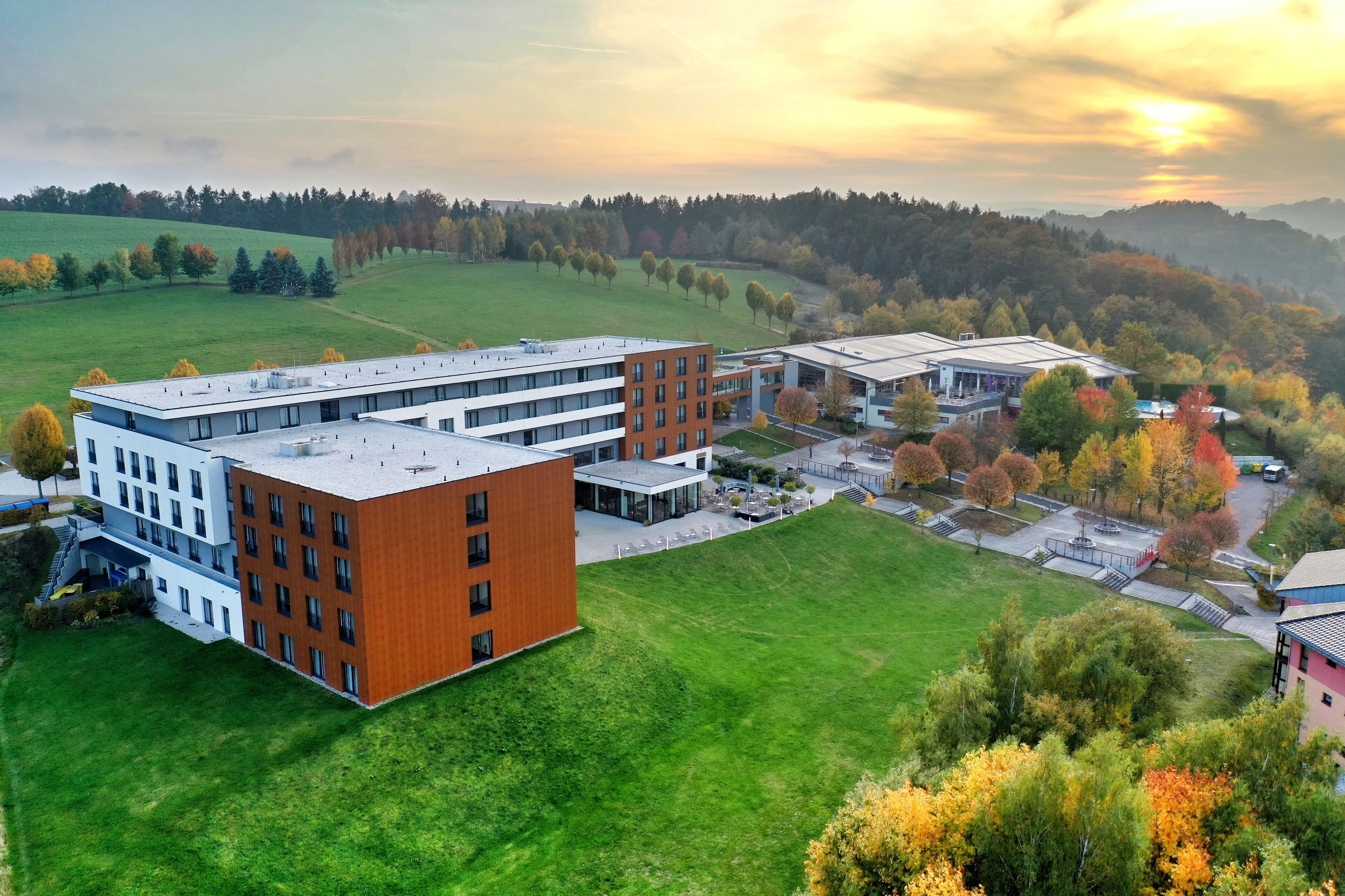 A modern building complex with multiple floors and a flat roof, surrounded by green fields and trees.