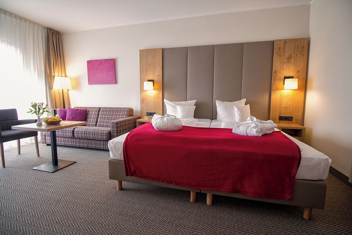 A hotel room with a large bed, a sofa, a small table with a fruit bowl, and two wall-mounted lamps.