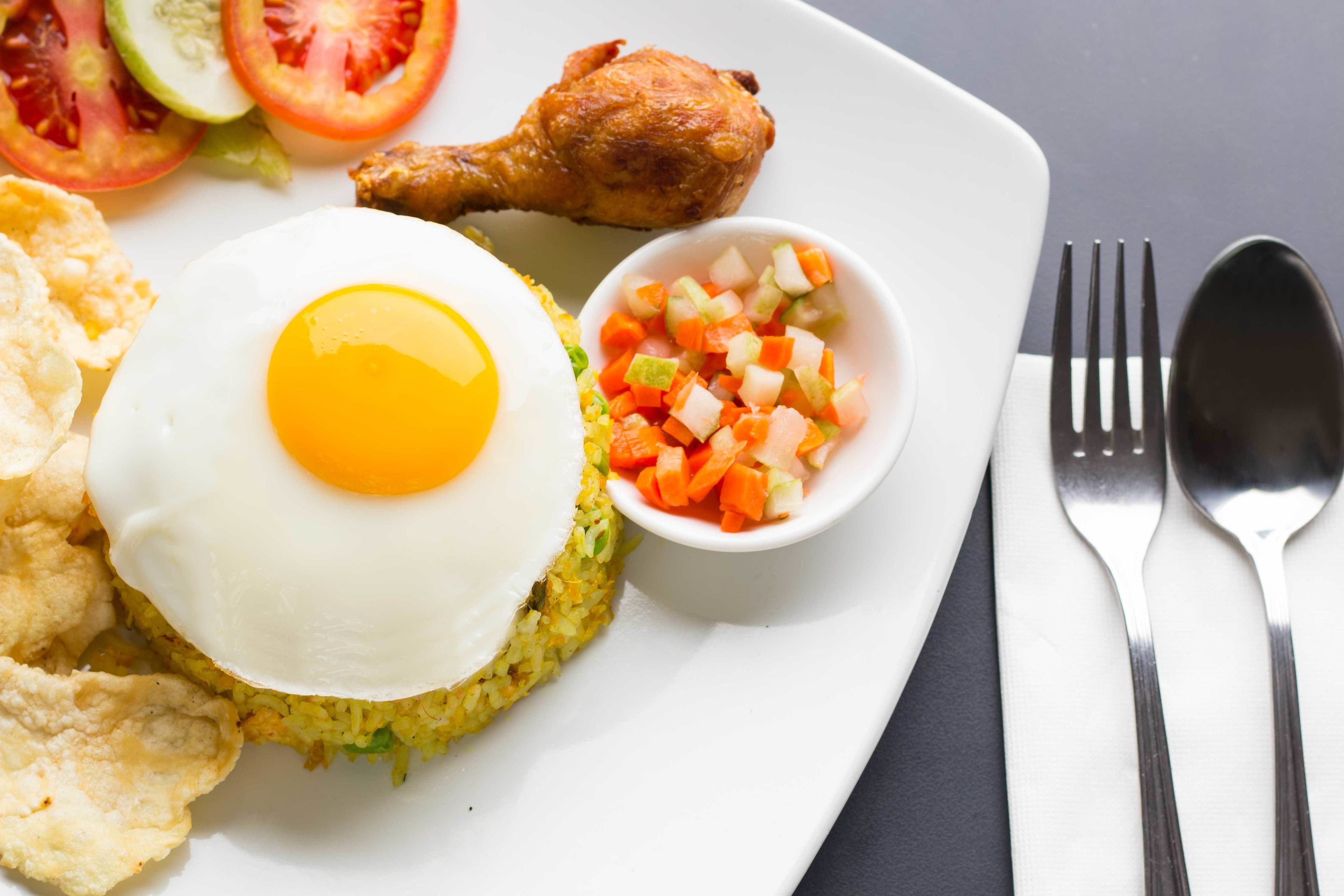 A plate with fried rice, a fried egg, a piece of fried chicken, a small bowl of diced vegetables, and a side of chips.