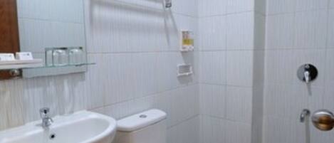 Casa de banho com lavatório branco, sanita e chuveiro. Existem prateleiras acima do lavatório e um porta-toalhas acima da sanita.