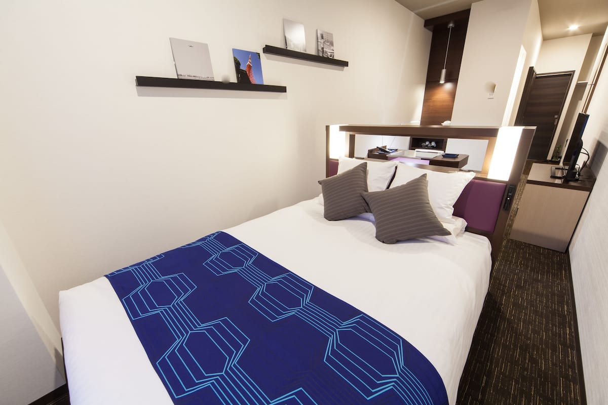 Quarto de hotel com cama, mesa de cabeceira, espelho e prateleira com livros e um quadro.