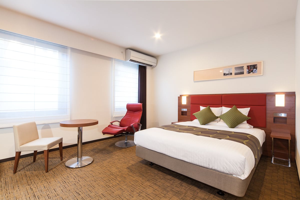 Quarto de hotel com uma cama grande, uma poltrona vermelha, uma mesa redonda pequena e uma janela com persianas.