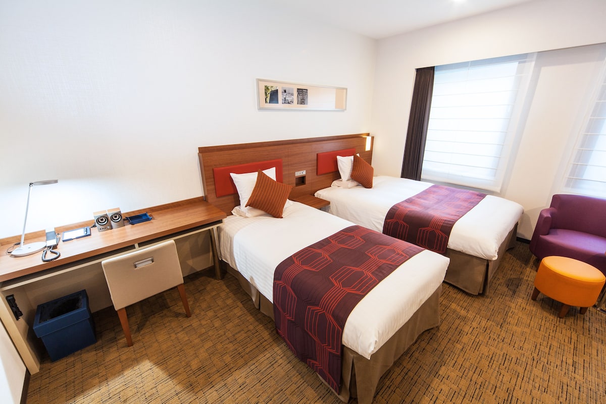 Quarto de hotel com duas camas, uma escrivaninha com computador, uma cadeira e uma janela com persianas.