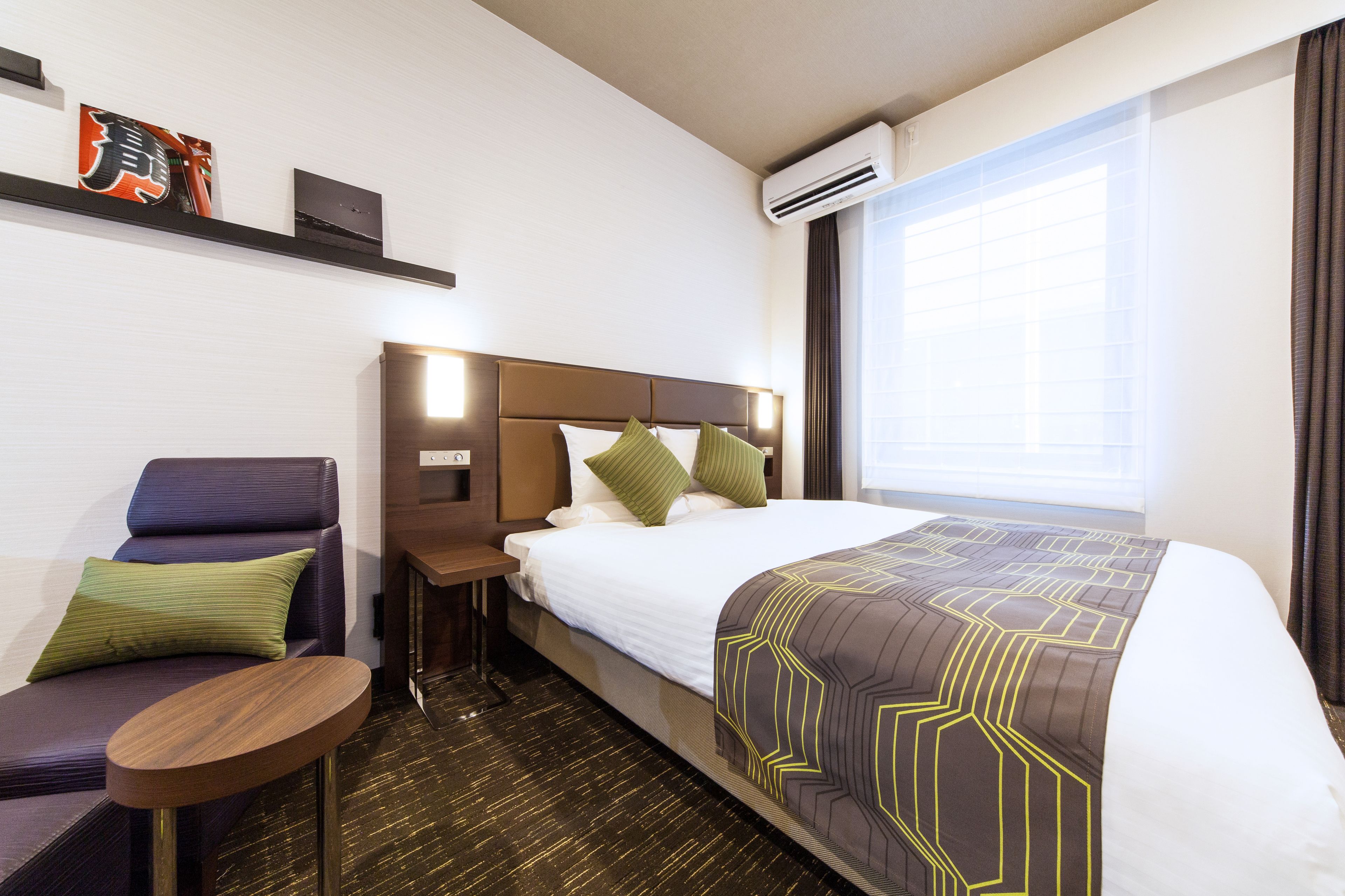 A hotel room with a bed, a sofa, a small table, a nightstand, and a wall-mounted air conditioner.
