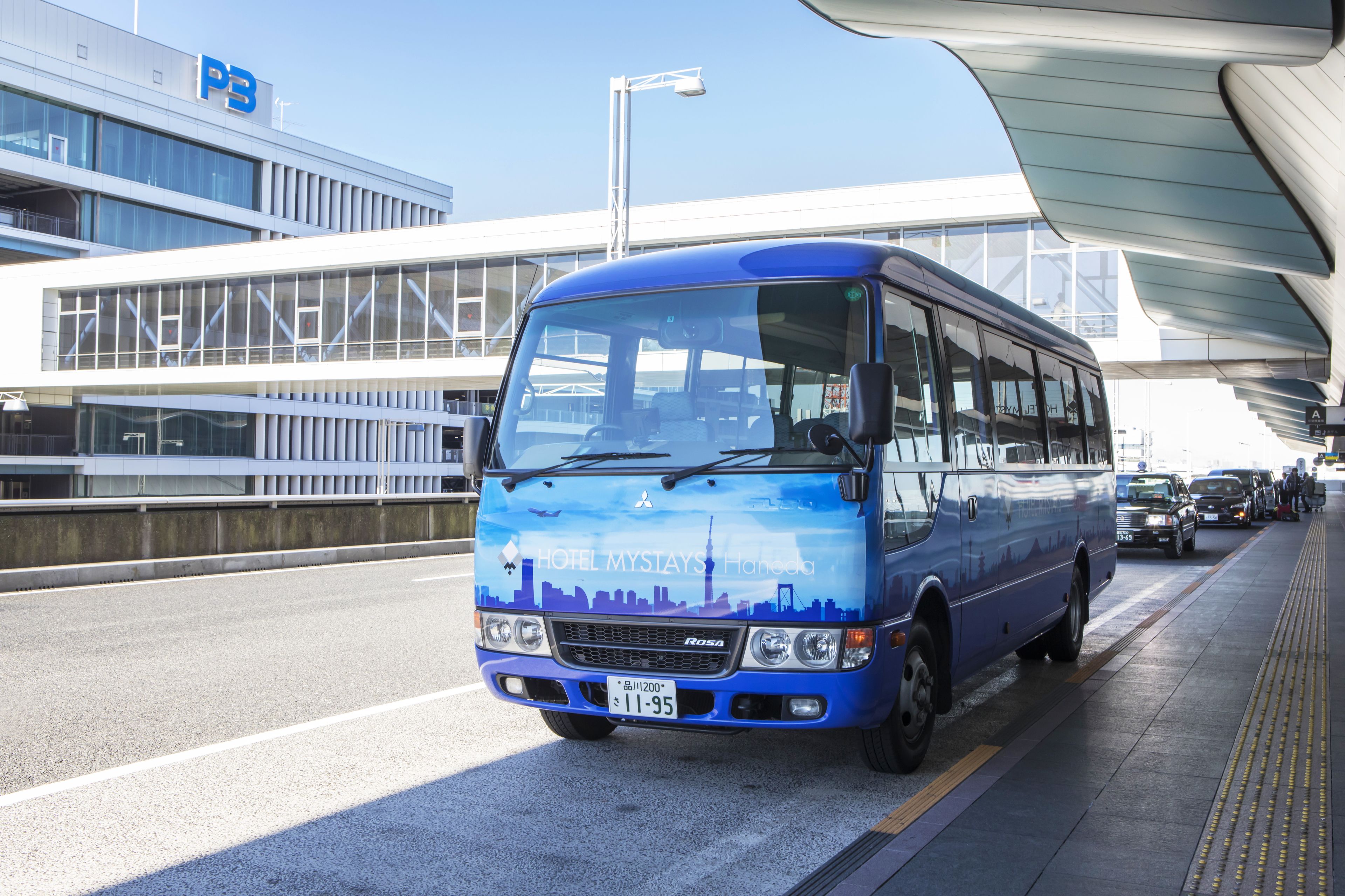 A blue bus with "HOTEL MYSTAYS Hohhot" on the front, parked on a road with a modern building in the background.