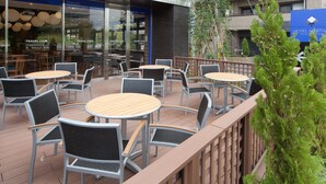 Un espace extérieur aménagé avec des tables et des chaises rondes, une clôture en bois et, en arrière-plan, une vue sur un café proposant des produits pour les voyageurs.