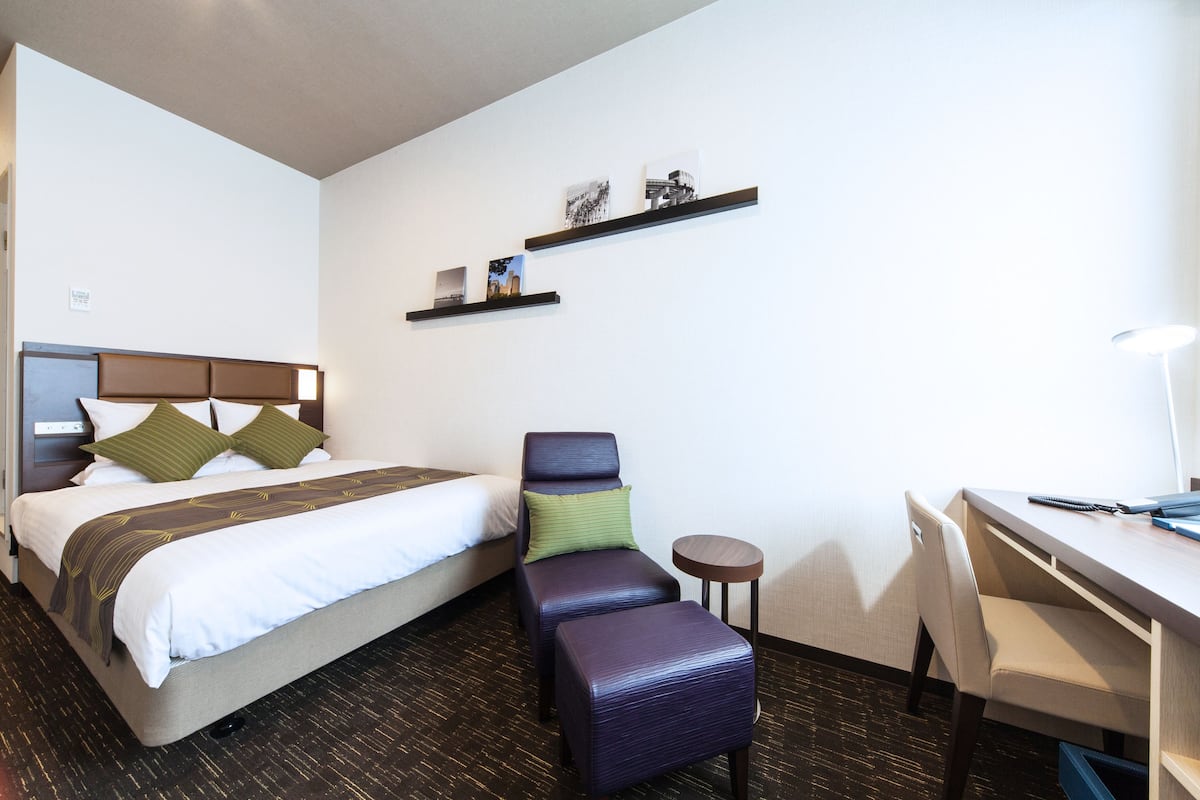 Quarto de hotel com cama, escrivaninha, cadeira e mesinha.