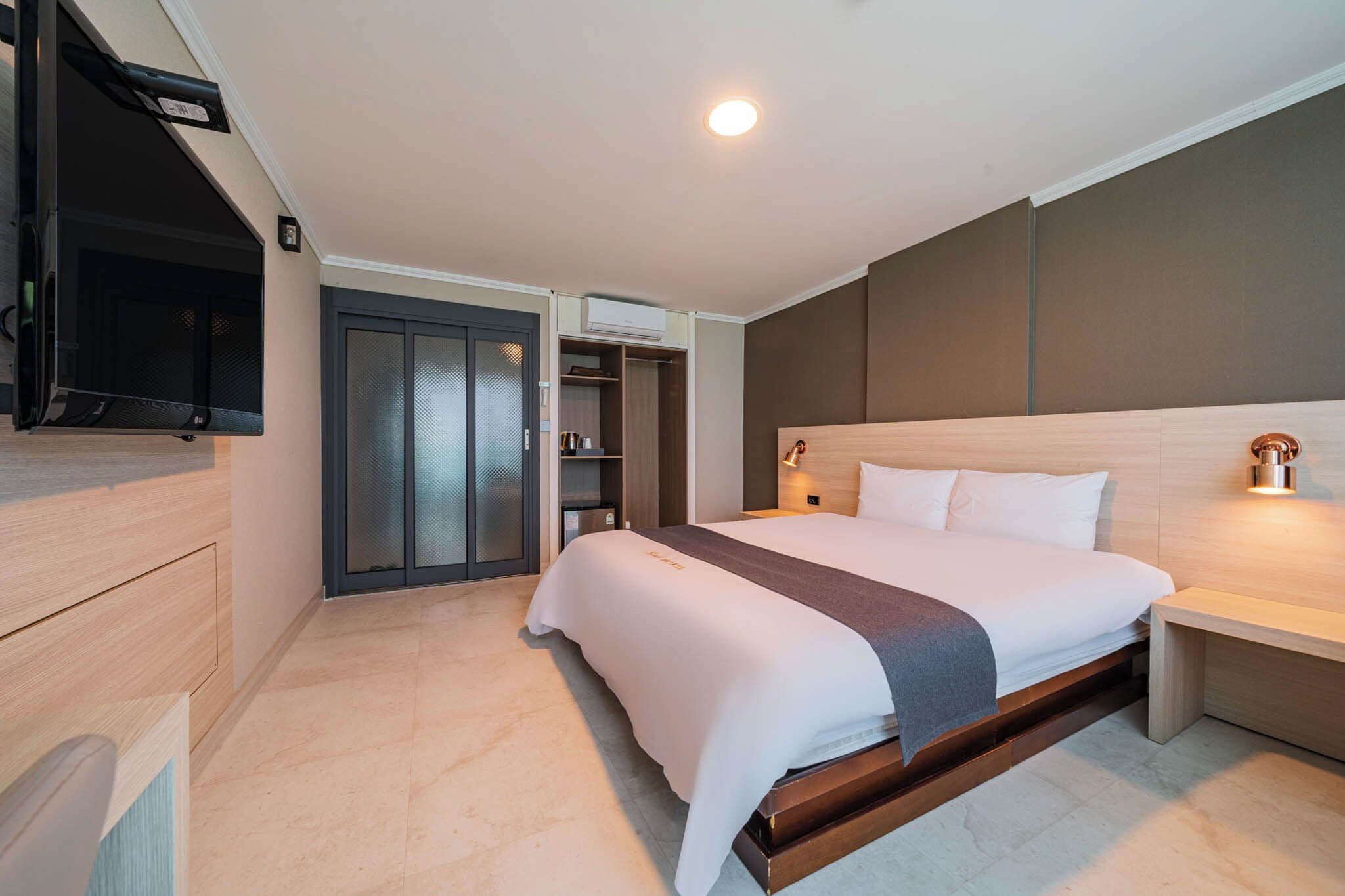 Ein modernes Hotelzimmer mit einem großen Bett, einem an der Wand montierten Fernseher, einem Kleiderschrank und einem Nachttisch mit Lampe.