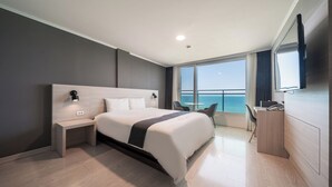 Une chambre d’hôtel moderne avec un grand lit, une table de chevet et un bureau avec une chaise. Une grande fenêtre offre une vue sur la mer.