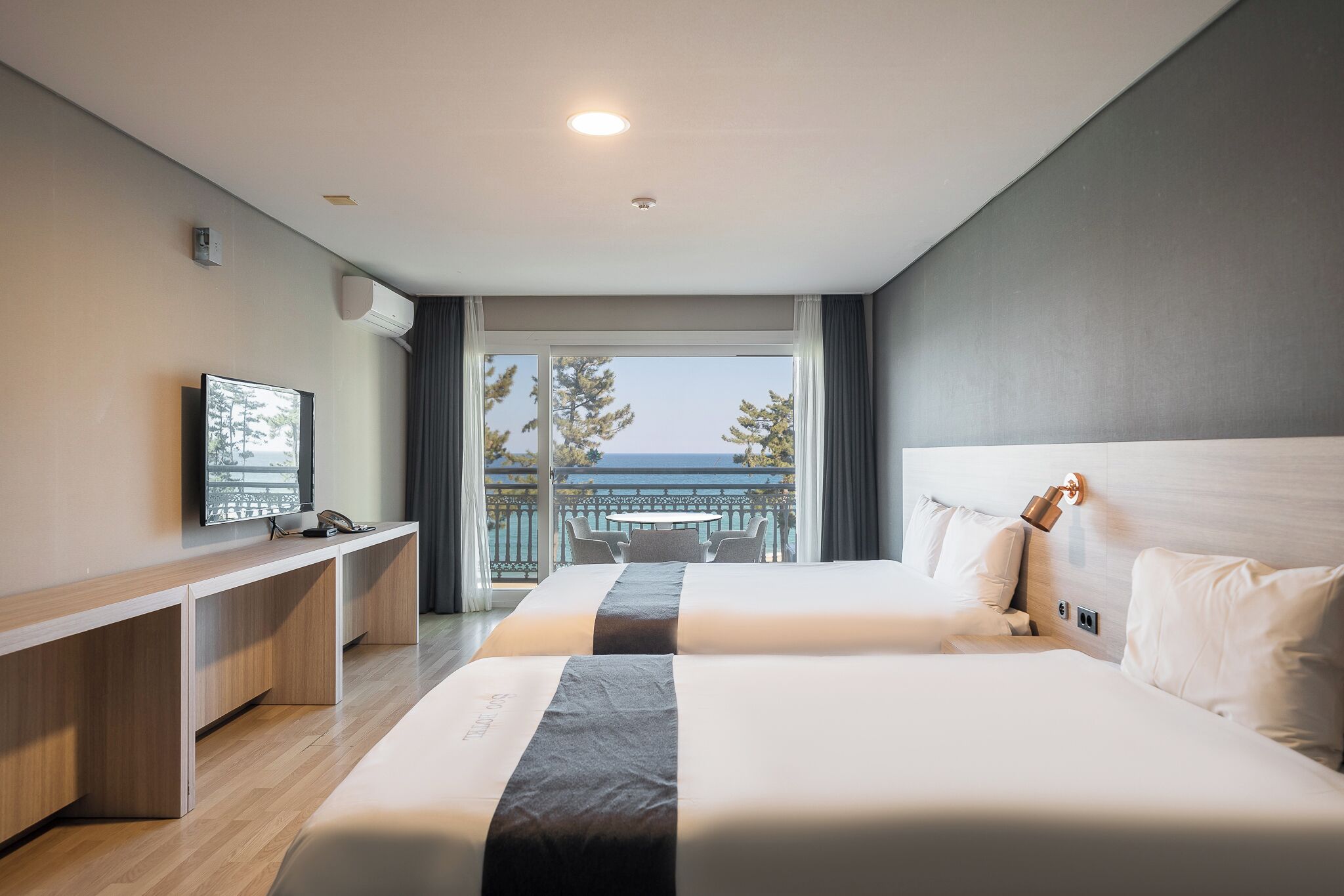 A hotel room with two beds, a desk, a TV, and a balcony with a view of the sea.