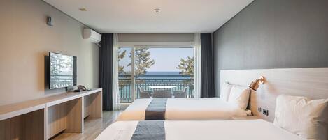 A hotel room with two beds, a desk, a TV, and a balcony with a view of the sea.