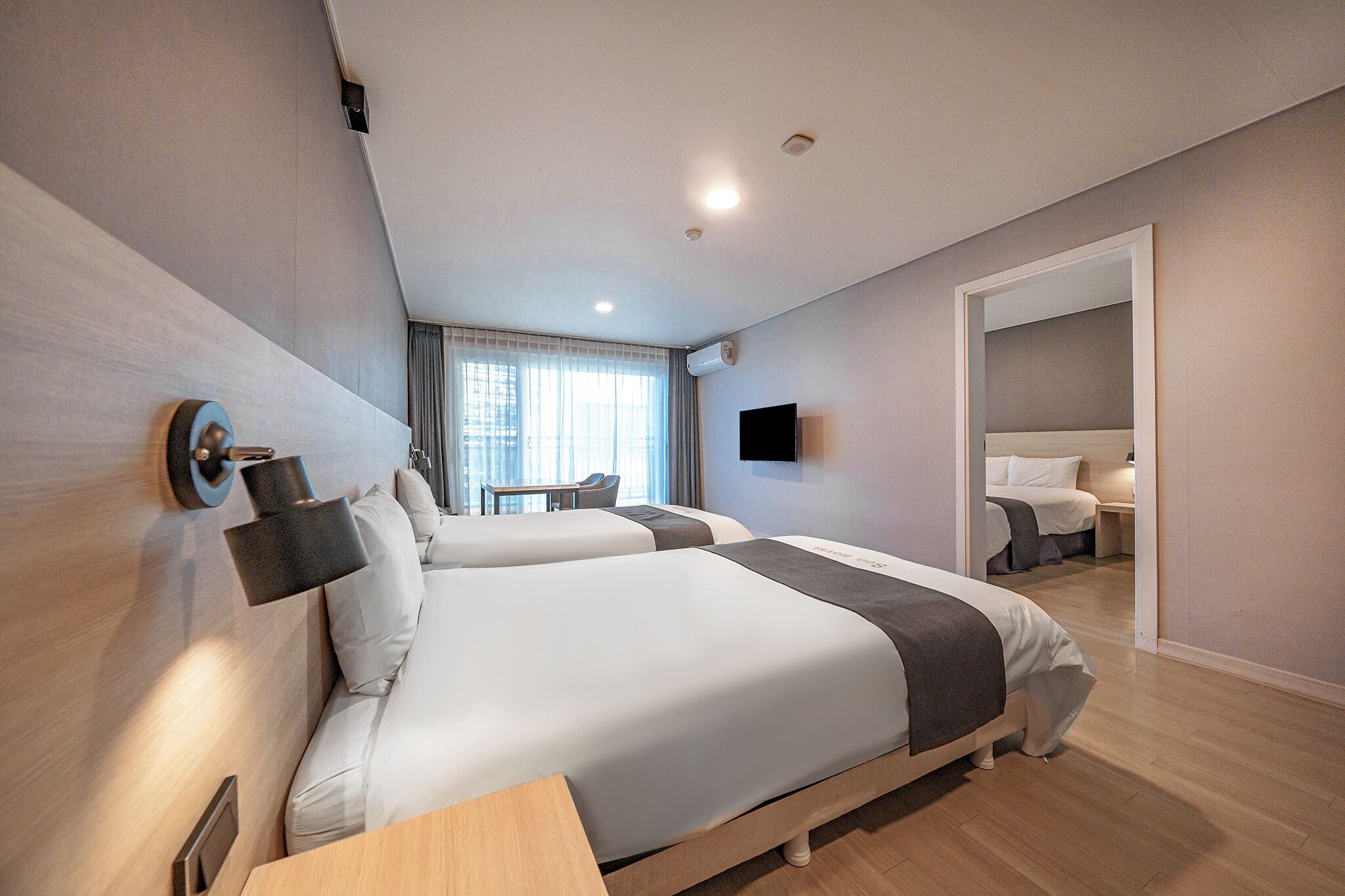 A modern hotel room with two beds, a desk, a flat-screen TV, and a large mirror.