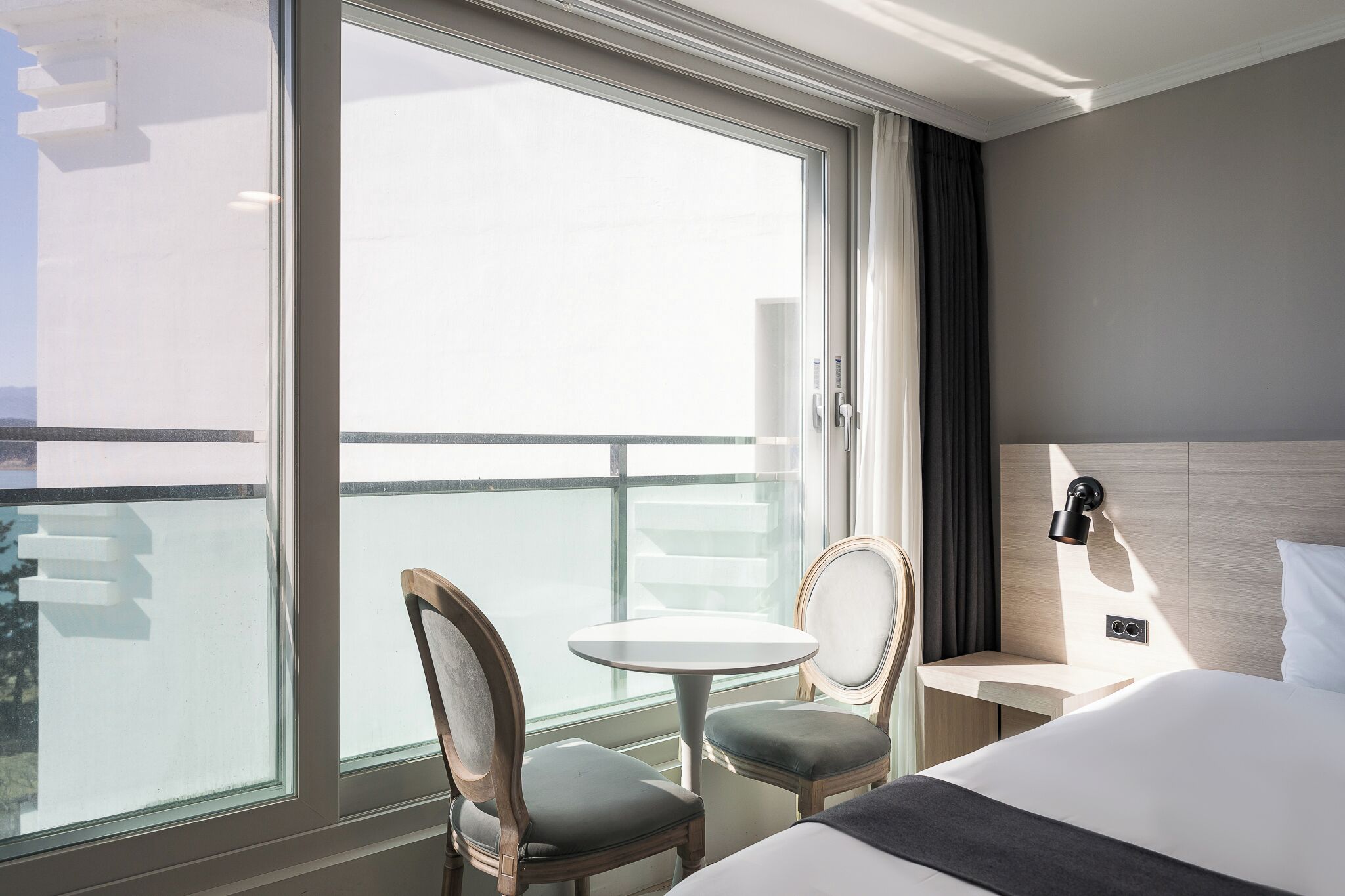 A hotel room with a large window, a bed, a bedside table, a lamp, and two chairs.