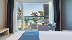 Une chambre d’hôtel avec un grand lit, une table ronde avec des chaises et un balcon offrant une vue sur la plage et les arbres.