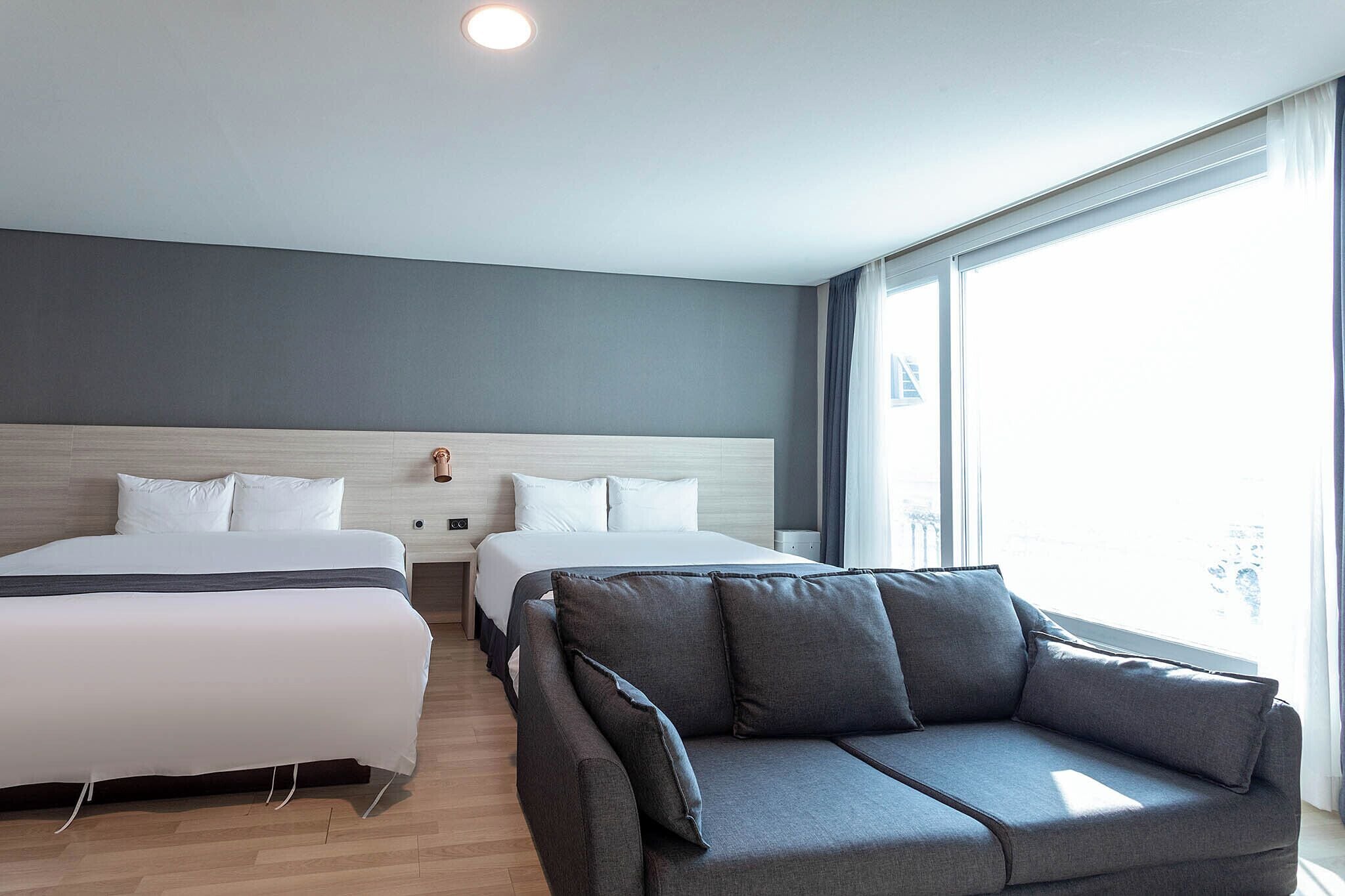 A modern hotel room with two beds, a grey sofa, and large windows offering a city view.