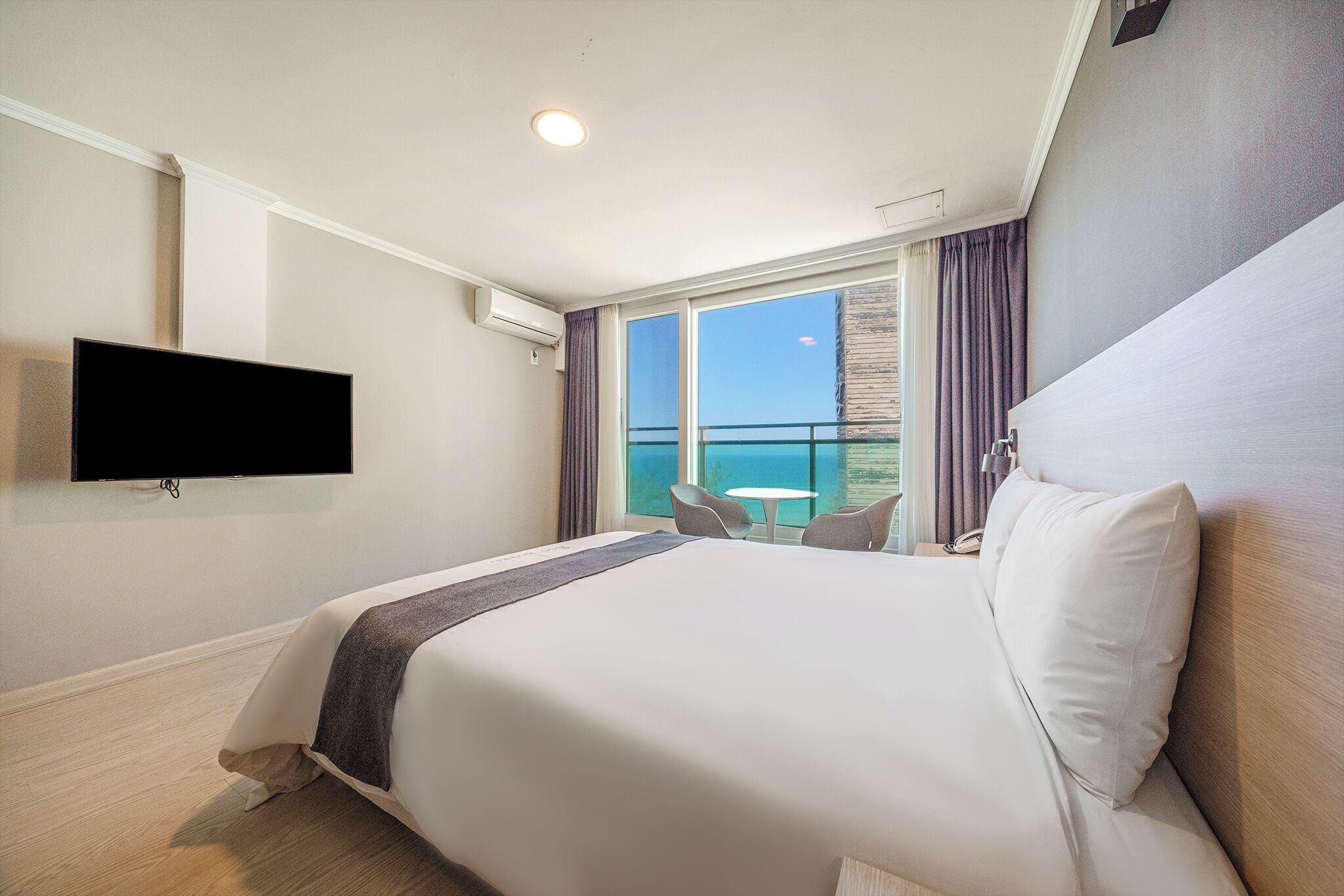 Een hotelkamer met een groot bed, een flatscreen televisie aan de muur, een klein rond tafeltje en uitzicht op de oceaan door de ramen.