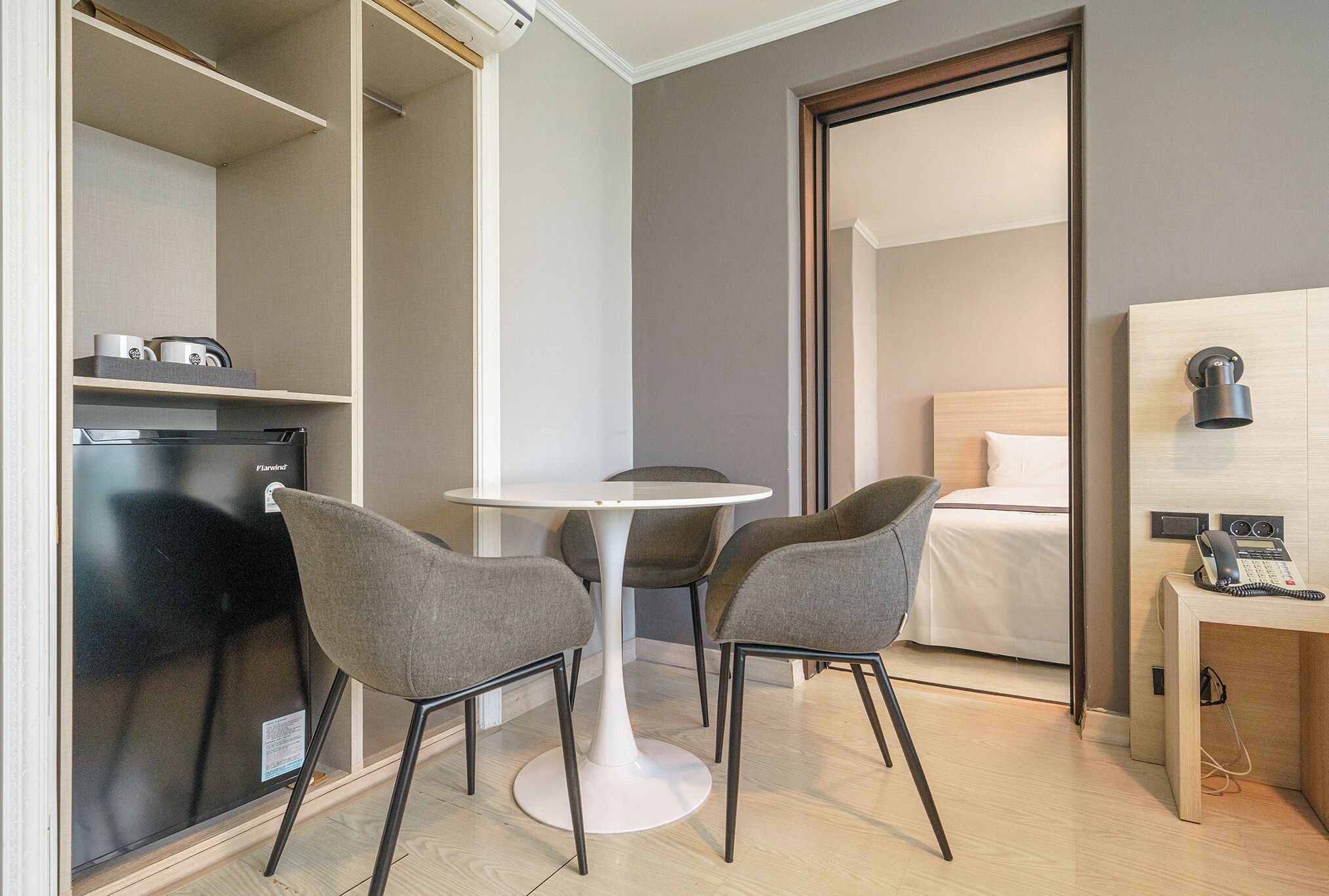 A compact hotel room with a bed, a desk with a telephone, a small table with two chairs, and a refrigerator.
