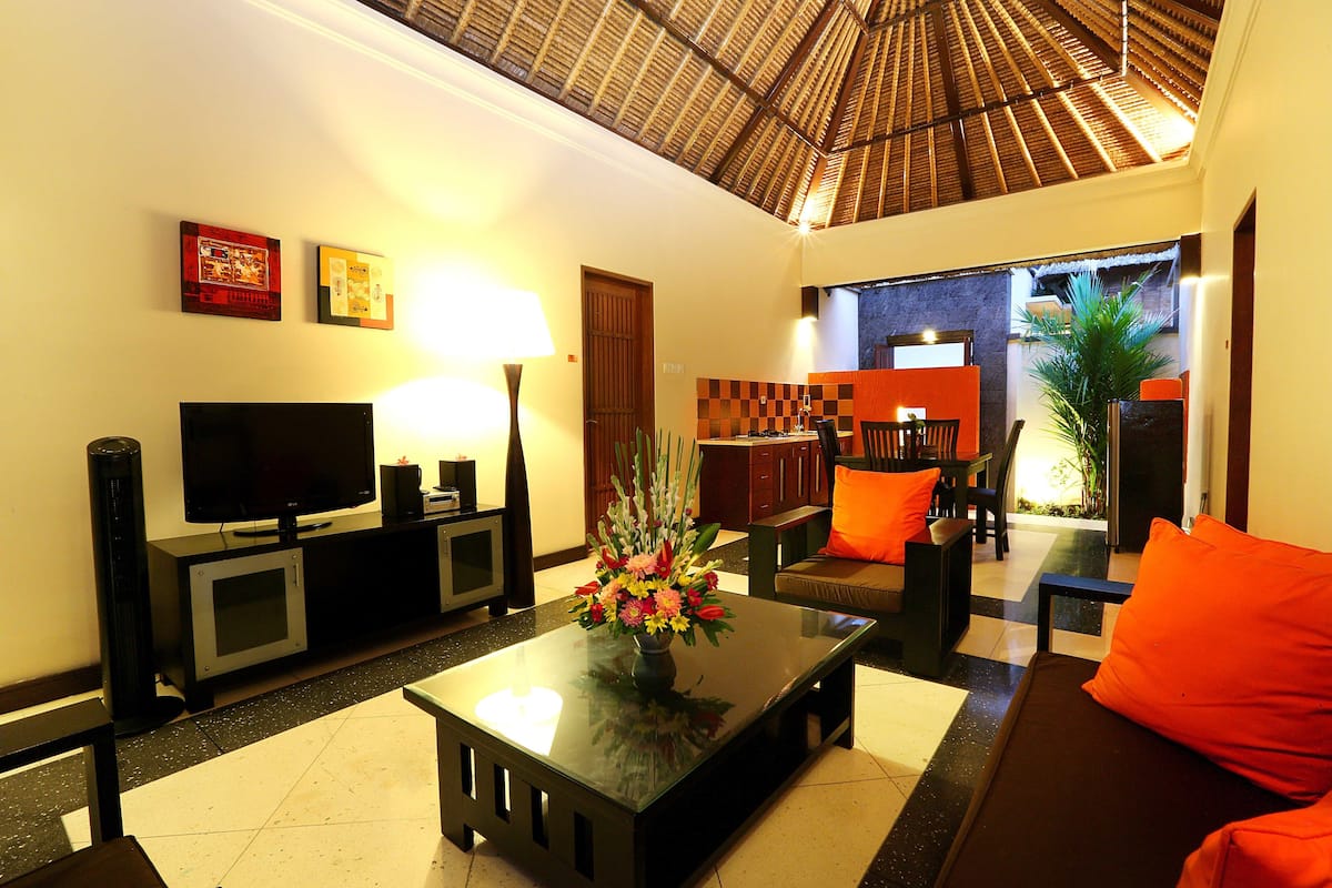 A modern living room with a flat-screen TV, a coffee table with a floral centerpiece, and a sofa with orange cushions.