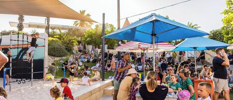 A crowded outdoor event with people sitting at tables under umbrellas, a playground with a climbing structure, and a sandy area.
