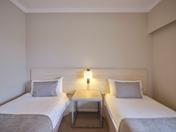Una habitación de hotel con dos camas individuales, una mesita de noche pequeña con una lámpara y una pared lisa.