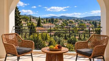 A balcony with wicker chairs and a table with fruit and a glass of wine, overlooking a scenic village.