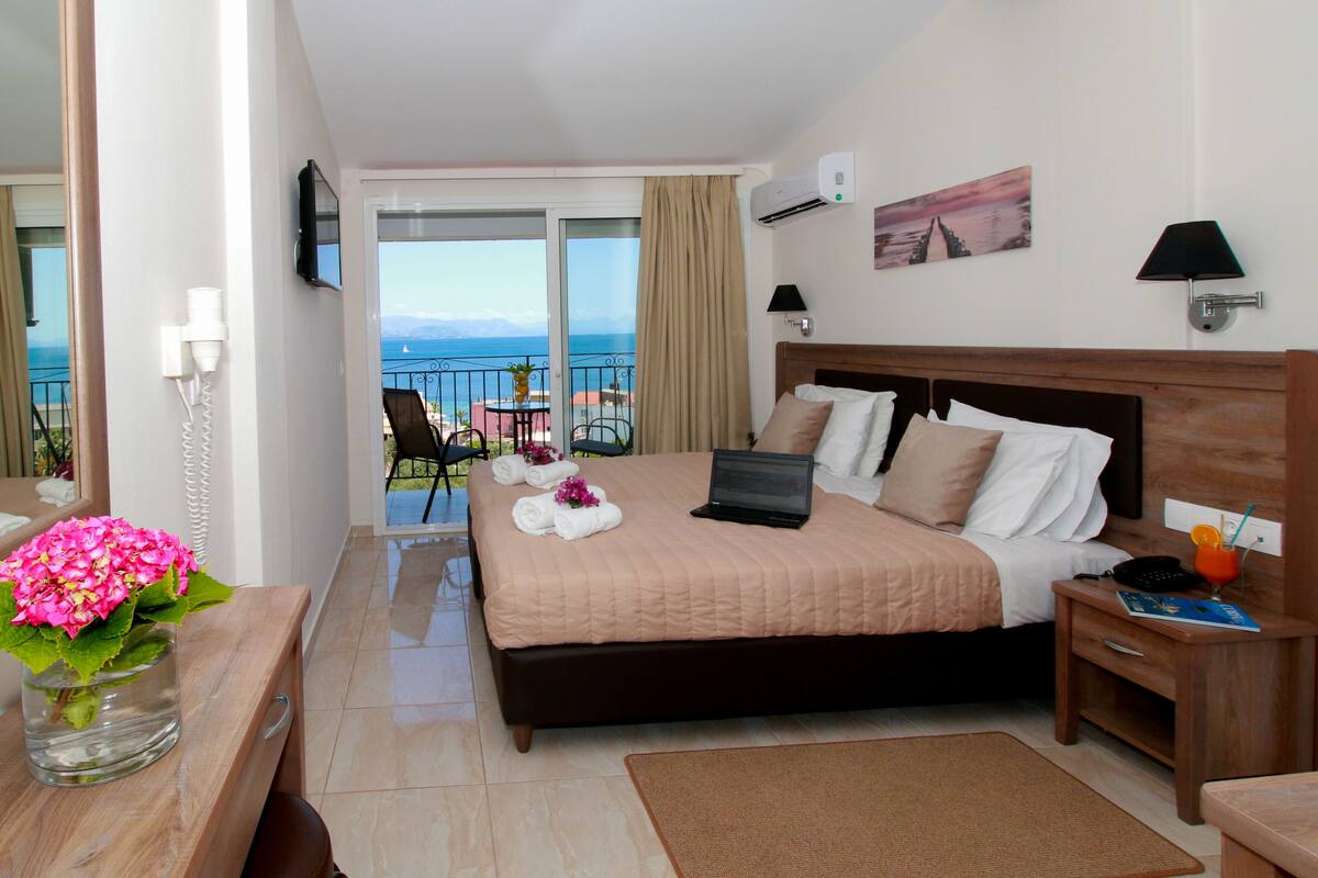Una camera d'hotel con un letto, un computer portatile, un comodino, una sedia e un balcone vista mare, oltre a un vaso con fiori.
