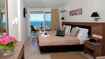 A hotel room with a bed, a laptop, a bedside table, a nightstand, a vase with flowers, a chair, and a balcony with a view of the sea.