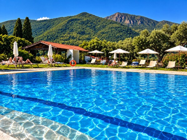 A swimming pool with clear blue water, surrounded by lounge chairs and umbrellas, with a mountainous backdrop. - Hotel Yannis Corfu (Corfu)