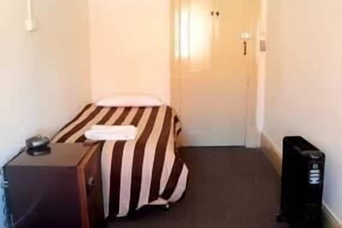 A small, narrow hotel room with a striped bed, a wooden dresser, and a black suitcase.
