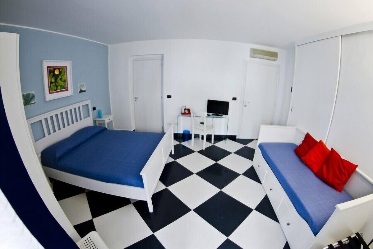 A room with a checkered floor, a bed with blue bedding, a sofa with red pillows, a desk with a computer, and a TV.