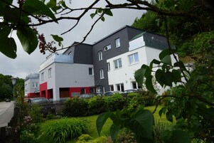 Un moderno edificio residencial de varios pisos con una combinación de fachadas blancas y grises, rodeado de abundante vegetación y un jardín bien cuidado.
