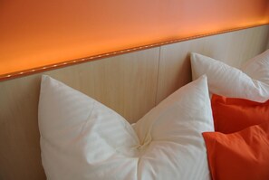 A bed with white and orange pillows against a wooden headboard with a warm light strip. - Pension Paradiesgartl (Amstetten)
