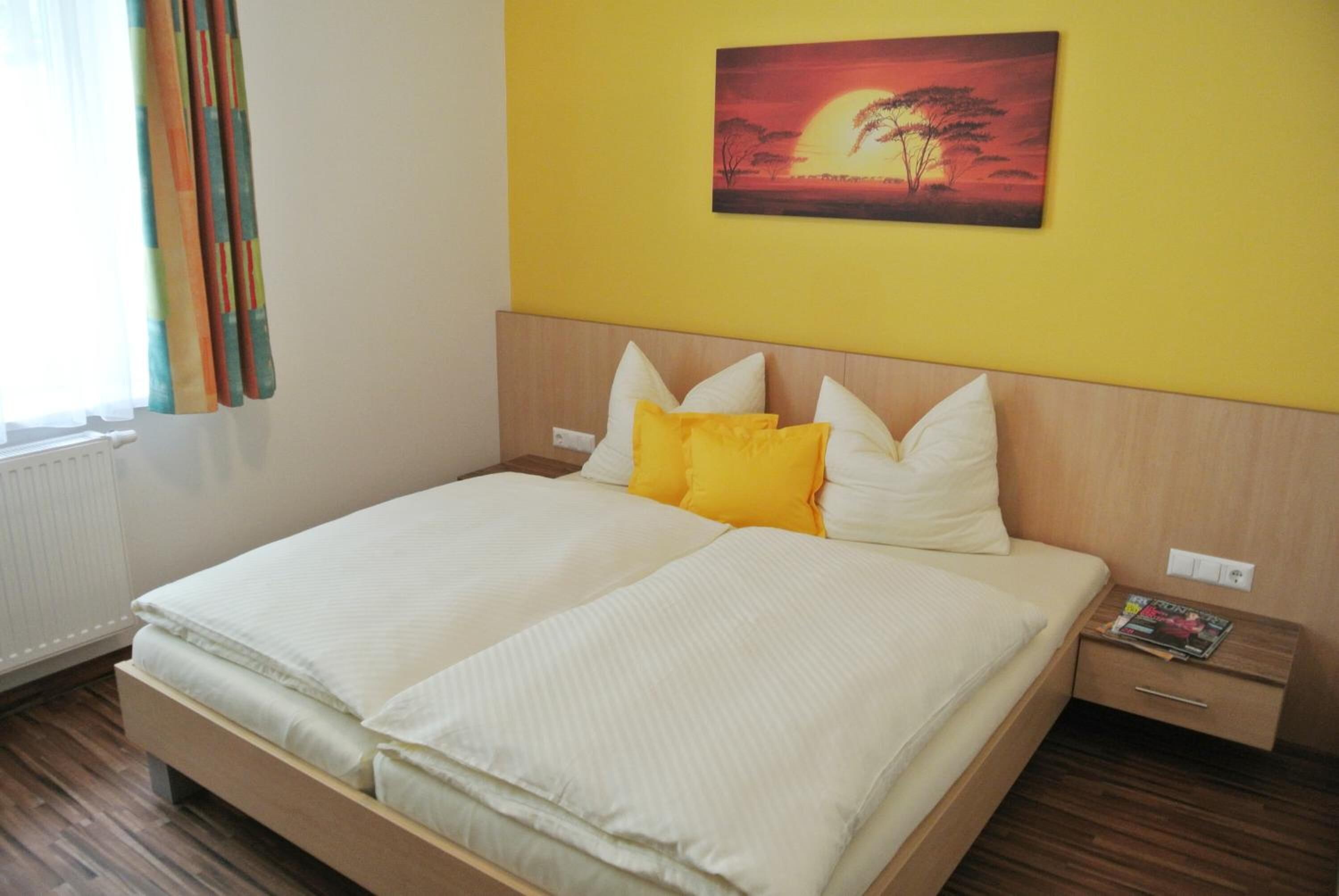A double bed with white and yellow bedding, a wooden headboard, and a nightstand with a magazine.